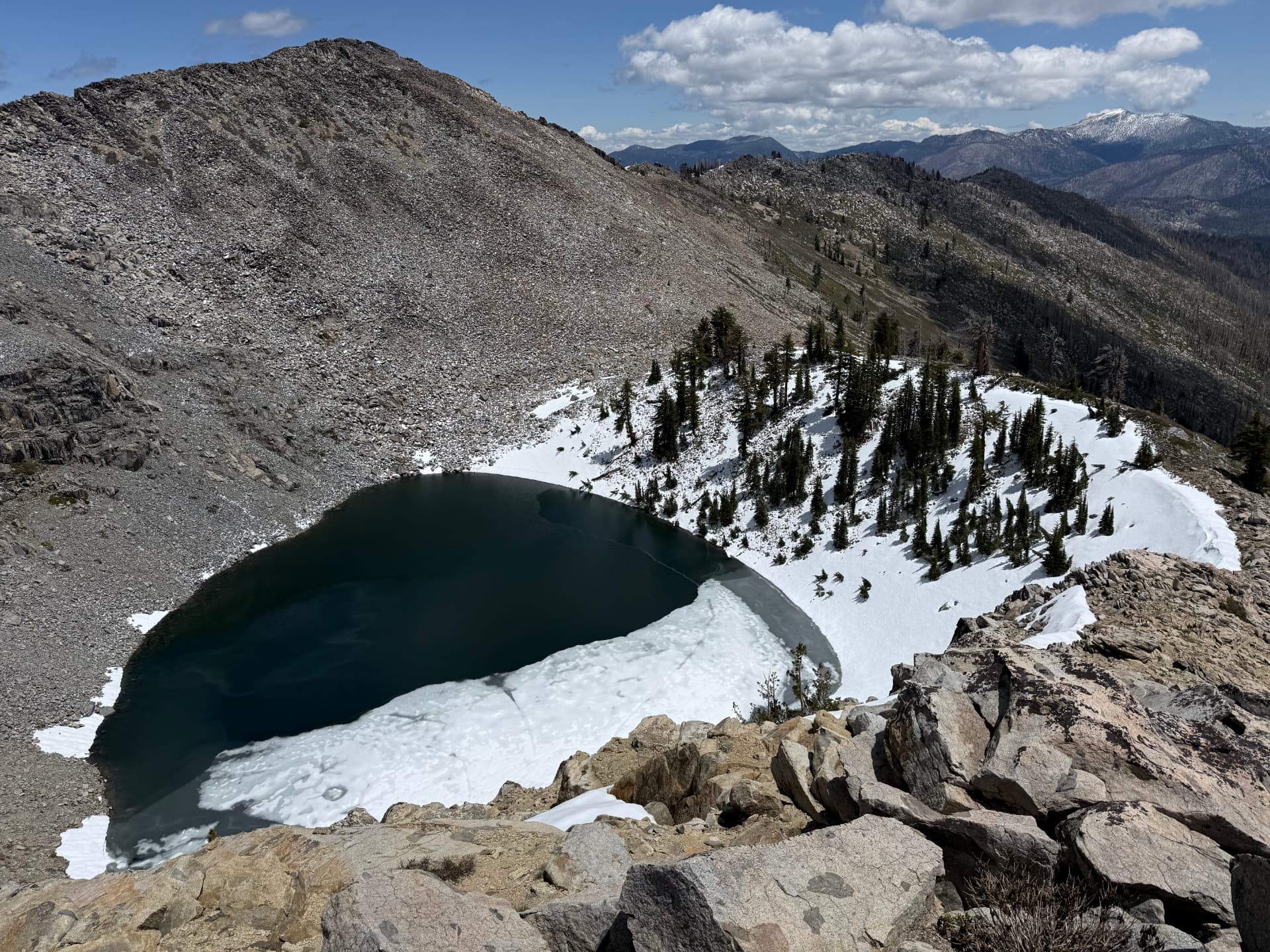 An aerial view of a dark alpine lake still holding ice along a rocky shoreline