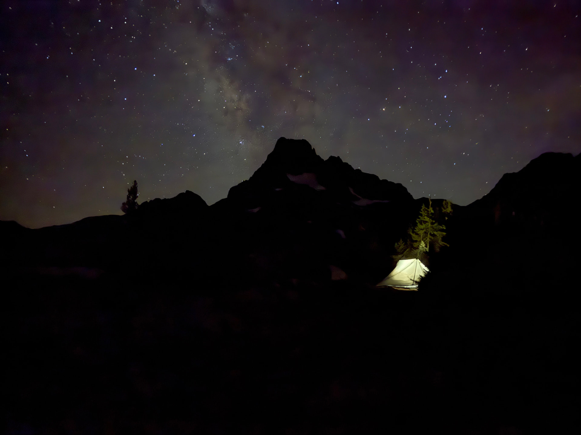 A glowing tent beneath the Milky Way with a dark mountain silhouette