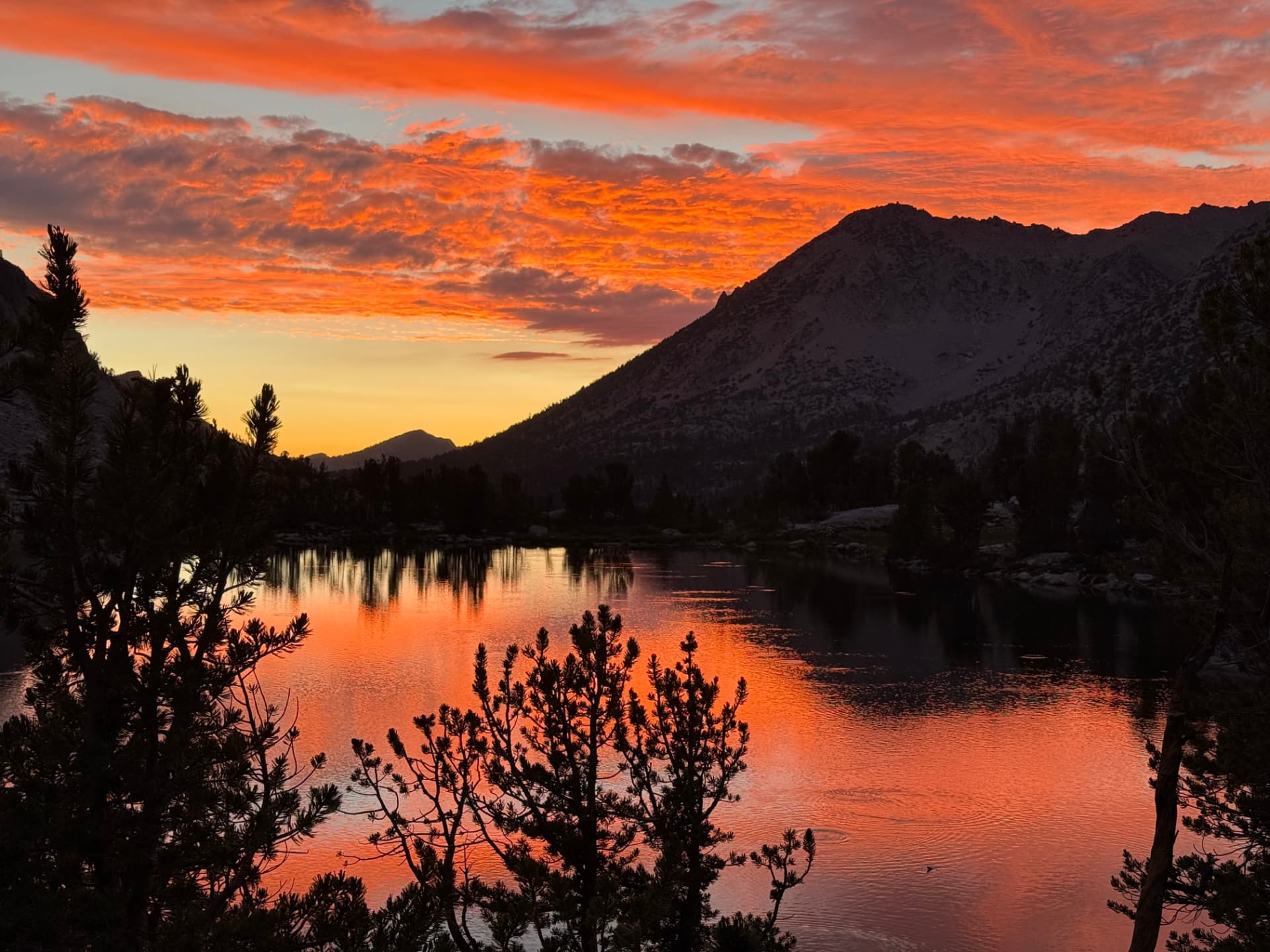 A fiery sunset sky reflected in a calm mountain lake with silhouetted peaks